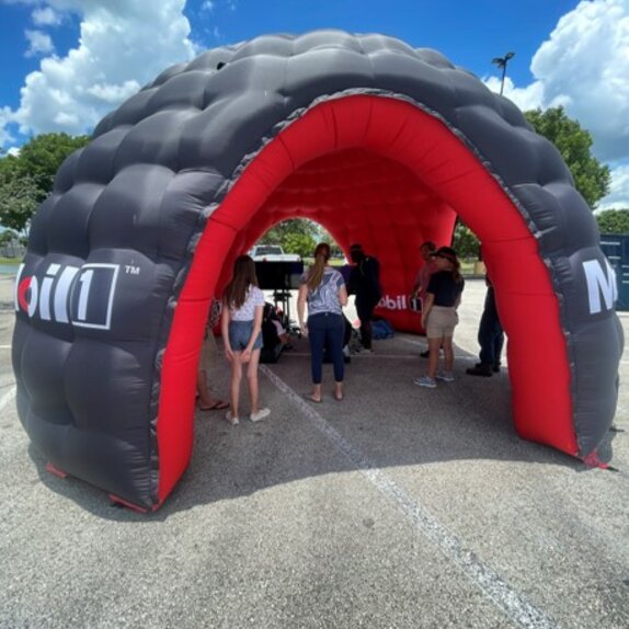 There is an inflatable advertising media of Mobil 1 in black and red on a square. Underneath it, there are some people.