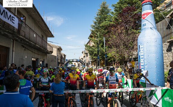 The picture shows many cyclists at the start of a race. On the right side there is a 6 m high inflatable advertising medium in the shape of a bottle.