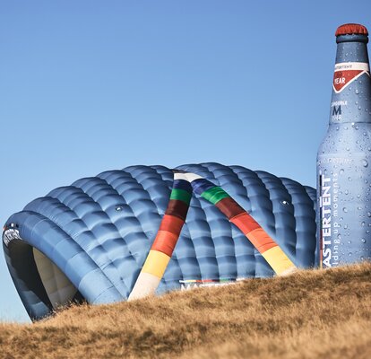 Zwei aufblasbare Werbeträger stehen auf einer Wiese nebeneinander: Modell "Kokon" und Modell "Bottle". Beide in den CI-Farben von MASTERTENT und mit einem Logo-Aufdruck.