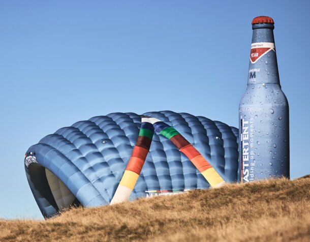 Two inflatable advertising media are located side by side on a meadow: model Kokon and model Bottle. Both in the CI colours of MASTERTENT and with a logo print.