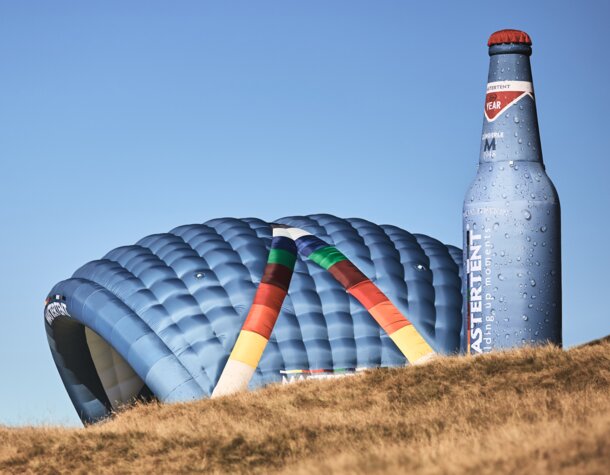 Two inflatable advertising media are located side by side on a meadow: model Kokon and model Bottle. Both in the CI colours of MASTERTENT and with a logo print.