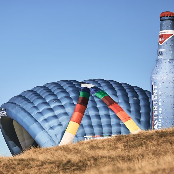 Two inflatable advertising media are located side by side on a meadow: model Kokon and model Bottle. Both in the CI colours of MASTERTENT and with a logo print.