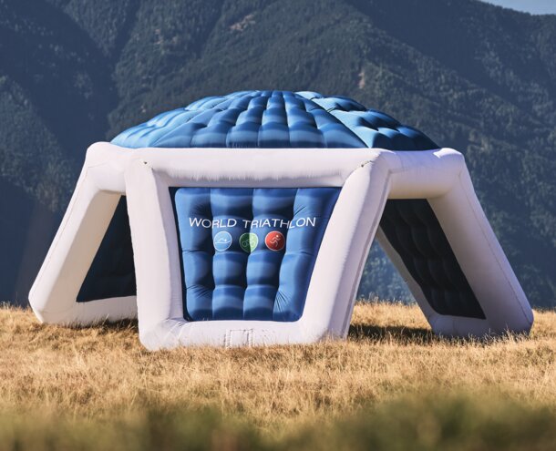 An inflatable advertising tent in blue and white with logo print "WORLD TRIATHLON" on a meadow in front of a mountain landscape.