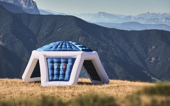 An inflatable advertising tent in blue and white with logo print "WORLD TRIATHLON" on a meadow in front of a mountain landscape.