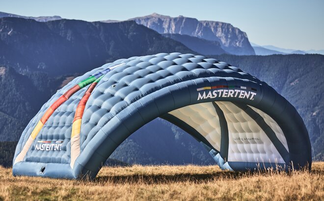 The inflatable advertising media Kokon in the CI colours of MASTERTENT with a logo print on the meadow. In the background of the arch-shaped advertising tent there is a mountain landscape.