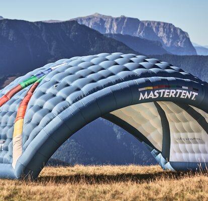 Der aufblasbare Werbeträger "Kokon" in den CI-Farben von MASTERTENT mit einem Logo-Aufdruck auf der Wiese. Im Hintergrund des Rundbogen-förmigen Werbezeltes befindet sich eine Berglandschaft.