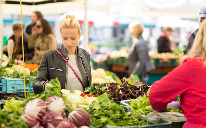 Eine Frau steht vor einem Marktstand und sucht sich Gemüse aus. 