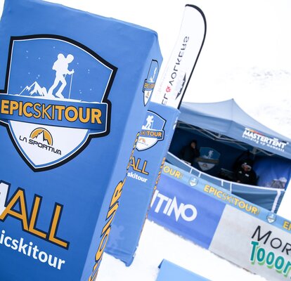 Im Vordergrund stehen blauen Pfosten mit dem Epic Ski Tour Logo und Absperrungen mit Werbung sind angebracht. Dahinter befindet sich ein blauer MASTERTENT Faltpavillon.