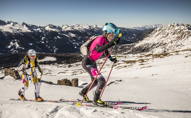 Una donna in tuta rosa sta salendo sulla montagna con gli sci. Viene seguita da un uomo in tuta giallo bianca.  