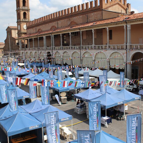 Blaue MASTERTENT Faltpavillons stehen in drei Reihen auf einem Stadtplatz. Über den Pavillons sind Fahnen von verschiedenen Nationen an einer Schnur aufgehängt.