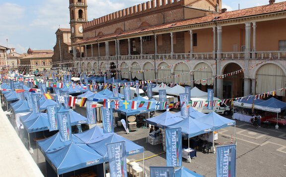 Blaue MASTERTENT Faltpavillons stehen in drei Reihen auf einem Stadtplatz. Über den Pavillons sind Fahnen von verschiedenen Nationen an einer Schnur aufgehängt.