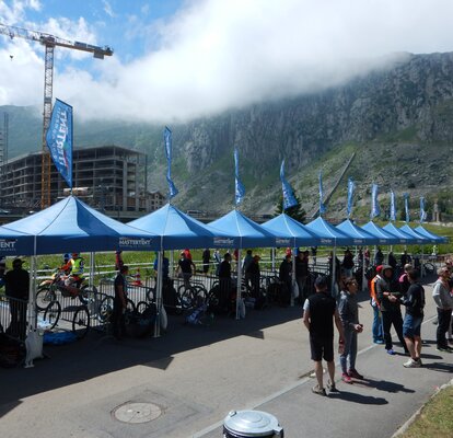 Molti gazebo pieghevoli MASTERTENT blu in una fila vicino la pista da gara. Sotto i gazebo pieghevoli ci sono delle bicilette e degli spettatori. 