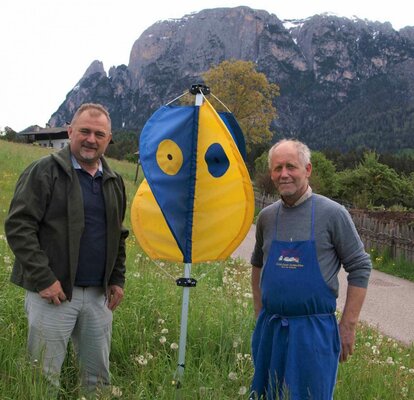 The fawn rescuer is placed like an umbrella in a meadow. He has blue and yellow flags. A hunter and a farmer stand next to it. 