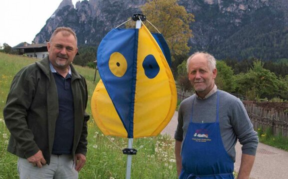 Kitzretter von Mastertent Der Kitzretter steht wie ein Regenschirm in einer Wiese. Er hat blaue und gelbe Fähnchen. Daneben stehen ein Jäger und ein Bauer.