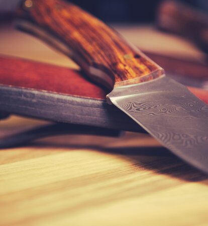 Das Messer liegt mit der Klinge nach vorne auf dem Tisch. Dahinter sieht man die Ledertasche des Messers.