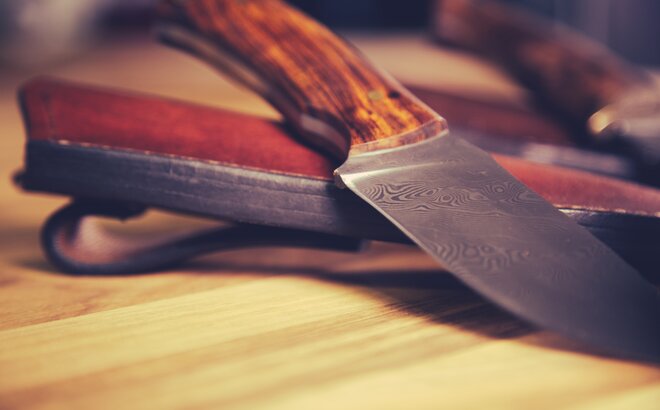 Das Messer liegt mit der Klinge nach vorne auf dem Tisch. Dahinter sieht man die Ledertasche des Messers.