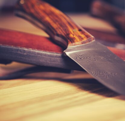 Das Messer liegt mit der Klinge nach vorne auf dem Tisch. Dahinter sieht man die Ledertasche des Messers.