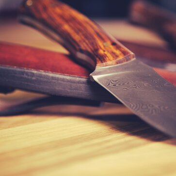 Das Messer liegt mit der Klinge nach vorne auf dem Tisch. Dahinter sieht man die Ledertasche des Messers.