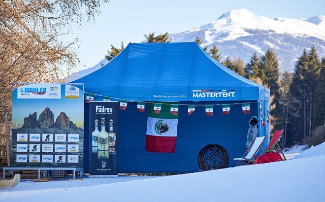 Der blaue Faltpavillon steht im Zielraum eines Skirennens. Er ist mit mexikanischen Fahnen dekoriert. Dahinter erstreckt sich ein schneebedeckter Berg.