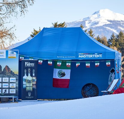 Der blaue Faltpavillon steht im Zielraum eines Skirennens. Er ist mit mexikanischen Fahnen dekoriert. Dahinter erstreckt sich ein schneebedeckter Berg.