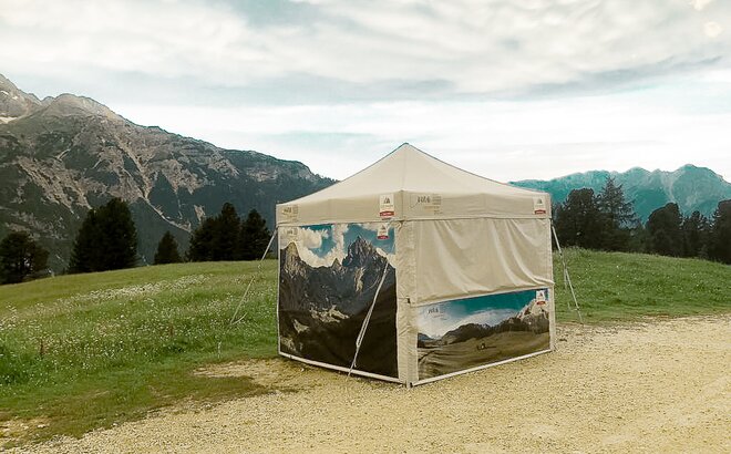 A gazebo from MASTERTENT, printed with pictures of lake Braies. It is located in the green meadow in front of mountains. 