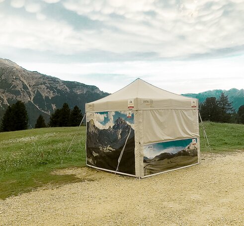 A gazebo from MASTERTENT, printed with pictures of lake Braies. It is located in the green meadow in front of mountains. 