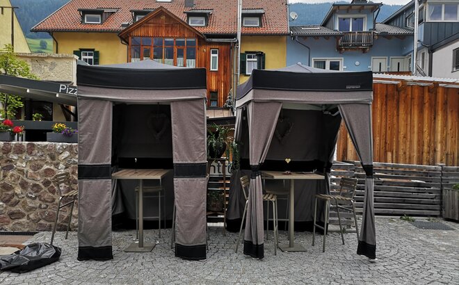 Zwei Lodenzelte stehen auf einer Terrasse. Beide Faltzelte haben eine geschlossene Faltzelt Seitenwand auf der Rückseite und Eckvorhänge vorne. 