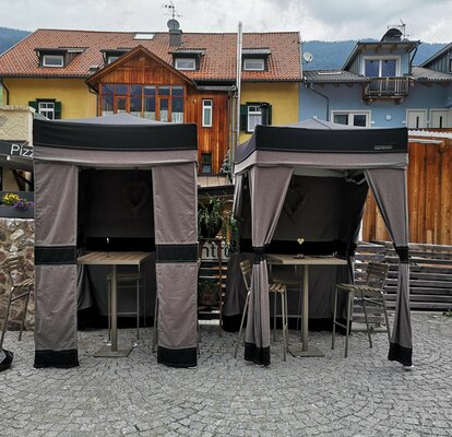 Two loden tents are standing on a terrace. Both gazebos have a closed side wall at the back and corner curtains at the front. 