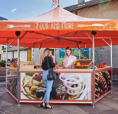 The saleswoman in the hexagonal vegetable stand is offering the customer a carrot to taste. The roof of the market stand is orange and printed with the words "FOOD AND MORE"; the panels, on the other hand, are printed with food all over.