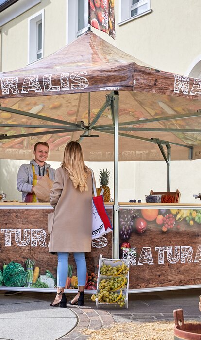 The seller in the fruit stand is serving his customers. He is handing over different kinds of fruit in a paper bag to a woman. The pavilion is printed with vegetables, fruit and the inscription "NATURALIS".