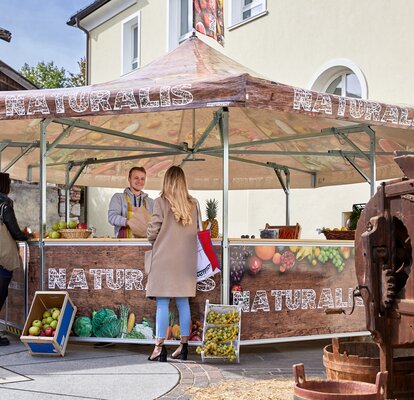 The seller in the fruit stand is serving his customers. He is handing over different kinds of fruit in a paper bag to a woman. The pavilion is printed with vegetables, fruit and the inscription "NATURALIS".