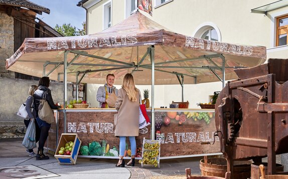 The seller in the fruit stand is serving his customers. He is handing over different kinds of fruit in a paper bag to a woman. The pavilion is printed with vegetables, fruit and the inscription "NATURALIS".