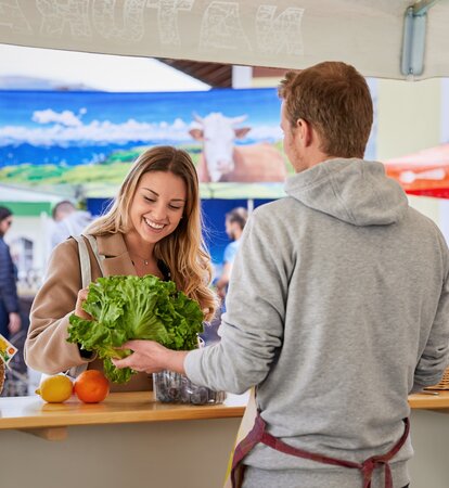 The seller in the fruit stall is informing the customer about his fresh fruit and vegetables. He is offering the woman a salad.