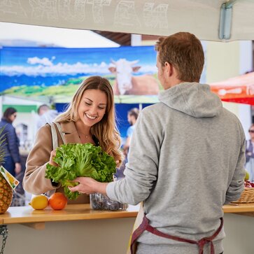 The seller in the fruit stall is informing the customer about his fresh fruit and vegetables. He is offering the woman a salad.