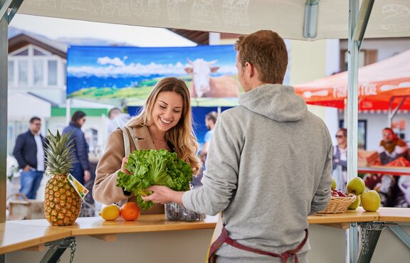 Der Verkäufer im Obststand klärt die Kundin über sein frisches Obst und Gemüse auf und hält ihr einen Salat hin.