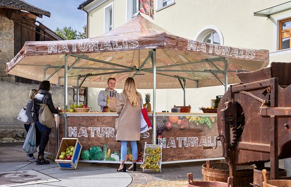 The seller in the fruit stand is serving his customers. He is handing over different kinds of fruit in a paper bag to a woman. The pavilion is printed with vegetables, fruit and the inscription "NATURALIS".