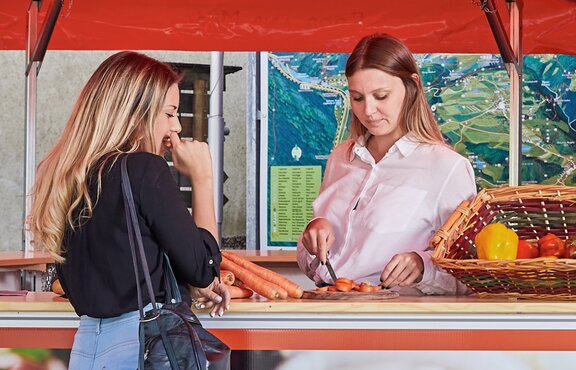 The saleswoman in the vegetable stand is cutting a carrot into thin slices. The customer in front of the pavilion is tasting a slice of carrot. 
