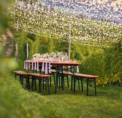 Stylisch dekorierte Bierzeltgarnituren mit weiß-grünen Blumen inmitten von Weinreben.