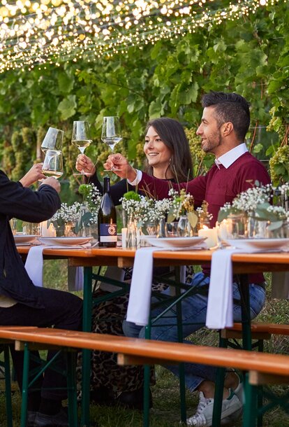 The beer tent set is decorated with white-green flower bouquets. The table and bench set is located n front of a grapevine. 4 people, including two couples, are sitting on the benches while toasting with wine glasses.