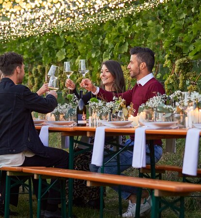 The beer tent set is decorated with white-green flower bouquets. The table and bench set is located n front of a grapevine. 4 people, including two couples, are sitting on the benches while toasting with wine glasses.