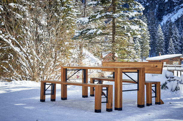 On a snow-covered terrace there is a wooden set consisting of a table, a bench with backrest and a bench without backrest. In the background are snow-covered trees.