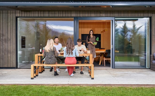 Four people are sitting on a Lago table and bench set which is located on the terrace of a modern residential building. A woman is carrying the salad from the kitchen to the garden. The group is having dinner in the garden. 