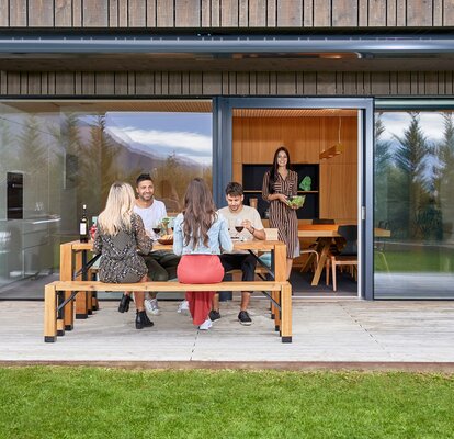 Four people are sitting on a Lago table and bench set which is located on the terrace of a modern residential building. A woman is carrying the salad from the kitchen to the garden. The group is having dinner in the garden. 