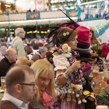 Souvenirverkäuferin mit Hüten, Stofftieren, Haarkränzen, Armbändern und herzförmigen Ansteckern auf dem Oktoberfest inmitten einer großen Menschenmenge, die auf Bierzeltgarnituren sitzt
