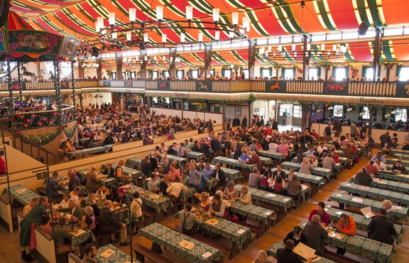 Das Marstall Festzelt auf dem Oktoberfest. Die Tische haben grüne Tischdecken. Die Gäste trinken und feiern im Zelt.
