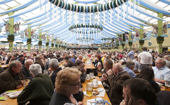Ochsenbraterei Festzelt von Innen. Am Dachboden hängen blau-weiße Schleifen. Im Zelt feiern Besucher auf den Garnituren.