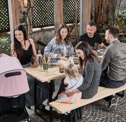 5 friends are sitting together on the "SitDown" beer table set on the terrace and dining. A woman is looking in the pram.