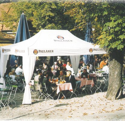 Ein weißer Paulaner Faltpavillon, unter dem Gäste eines Restaurant sitzen und etwas konsumieren.