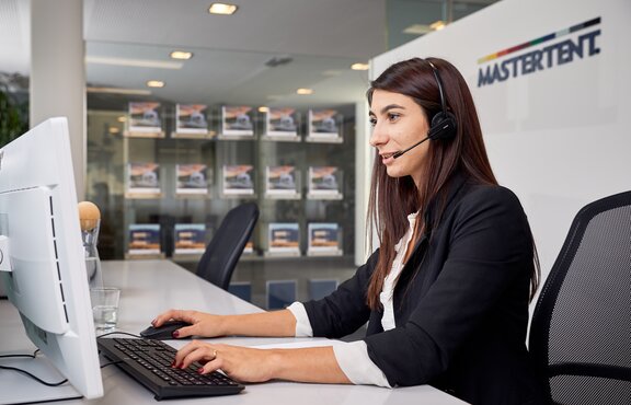 The employee is sitting with her head set in front of the computer while speaking to a client at the phone. 