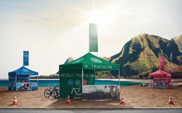 Drei Faltpavillons stehen auf dem Strand bei einem Triathlon. Die Faltpavillons sind grün, blau und rot. Alle drei haben eine Fahne und sind bedruckt. 